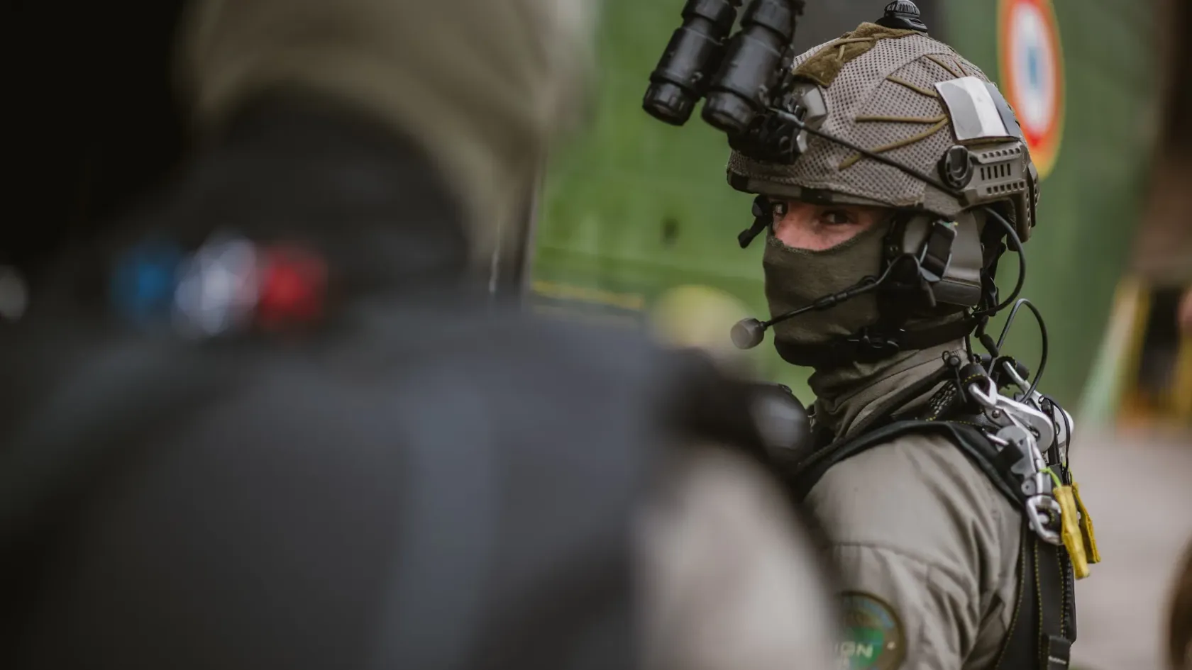 Operator with a helmet-mounted optic and headset microphone, geared for radio comms.