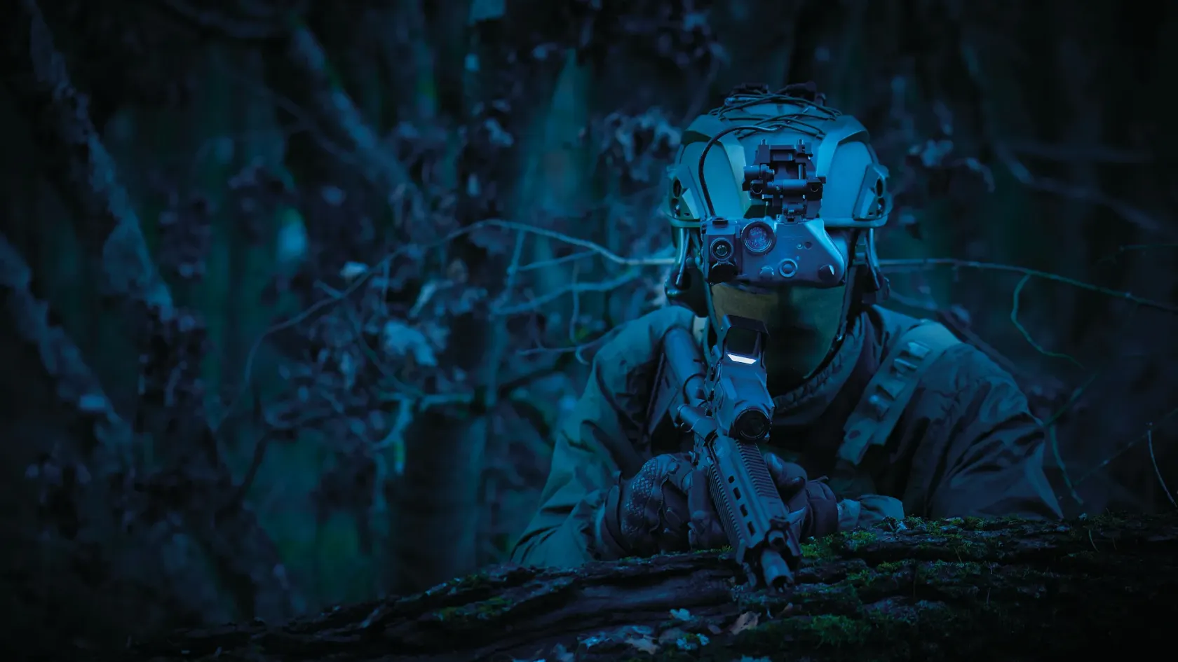 Operator at night sighting down a weapon, lit only by ambient light, in a green-blue night cast.